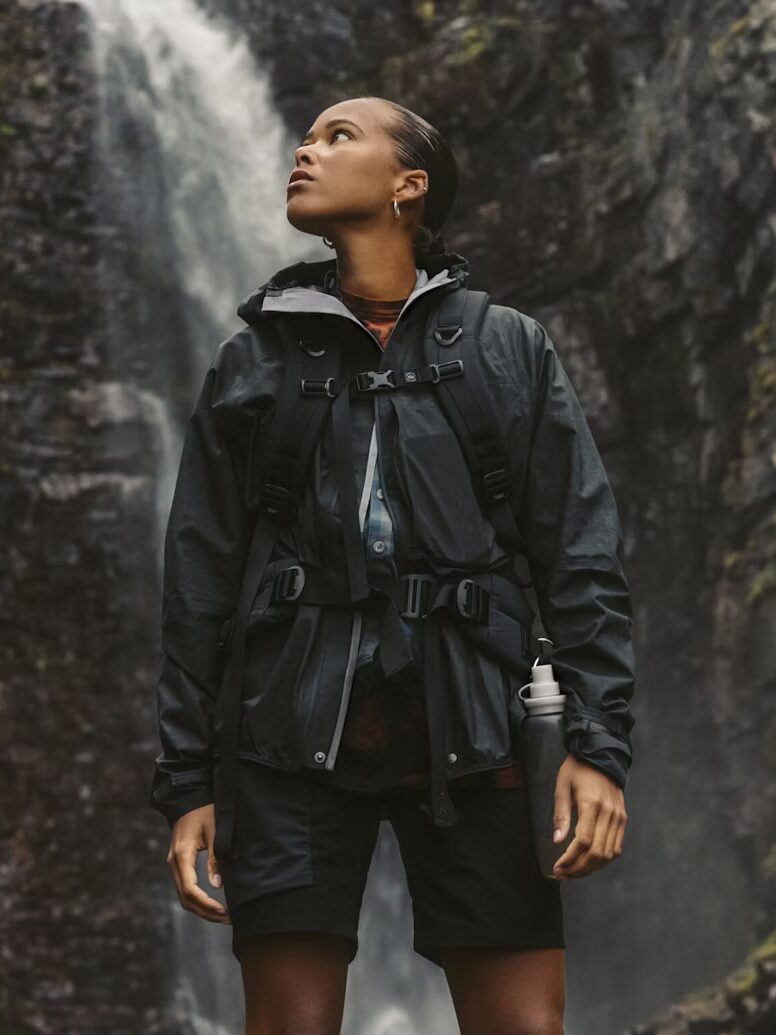 A person in a black waterproof raincoat and shorts stands outdoors near a rocky waterfall, looking upward with a water bottle in hand.