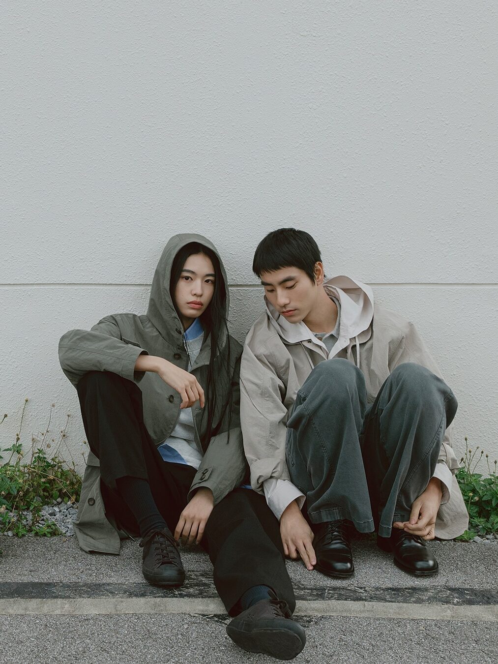 Two people sit on the ground against a light-colored wall, both in neutral-toned jackets—one wearing a waterproof raincoat with the hood up, the other looking down. Both have dark pants and black shoes.