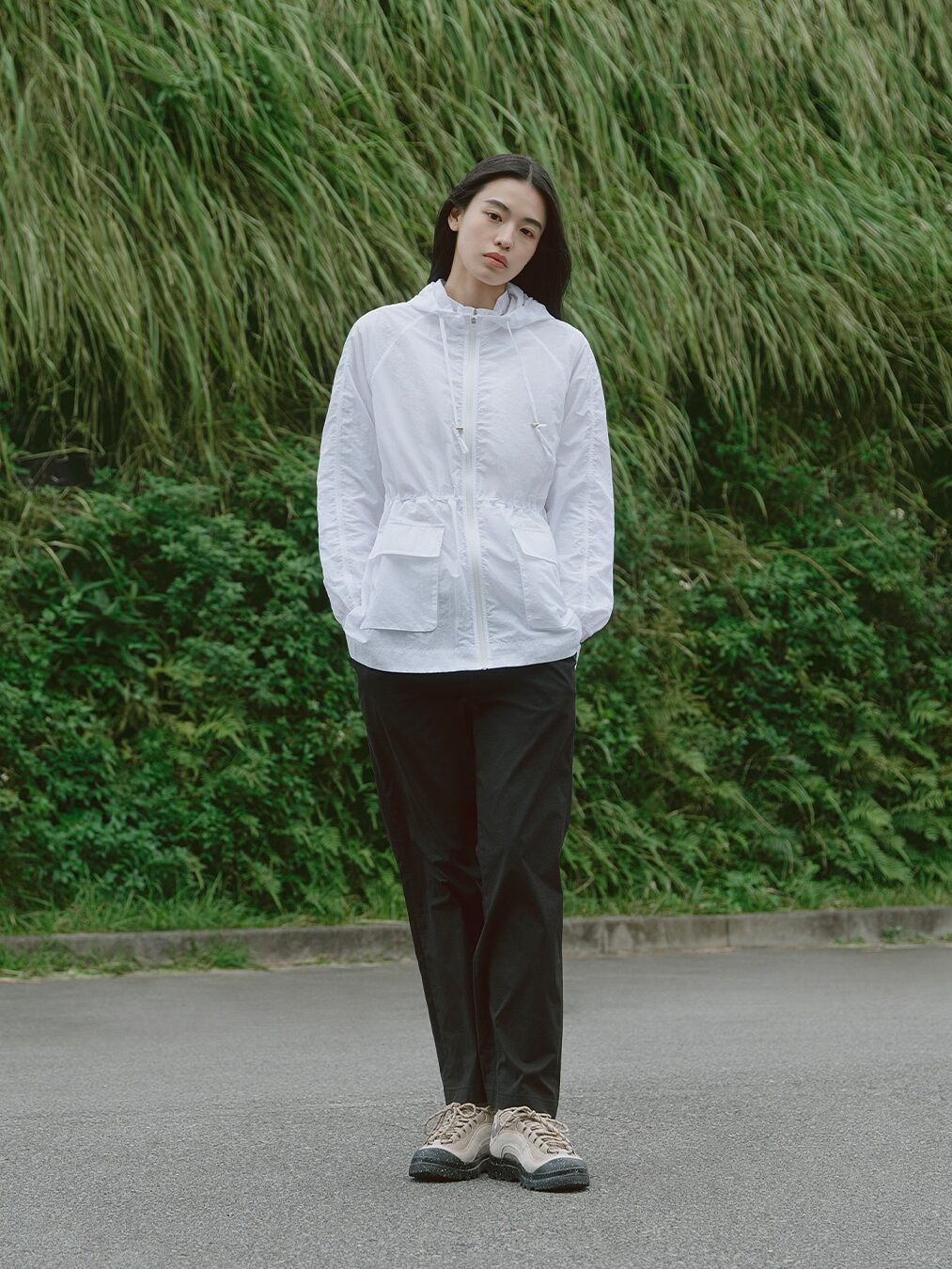A person wearing a white waterproof raincoat, black pants, and beige shoes stands on a paved surface with tall green grass in the background.