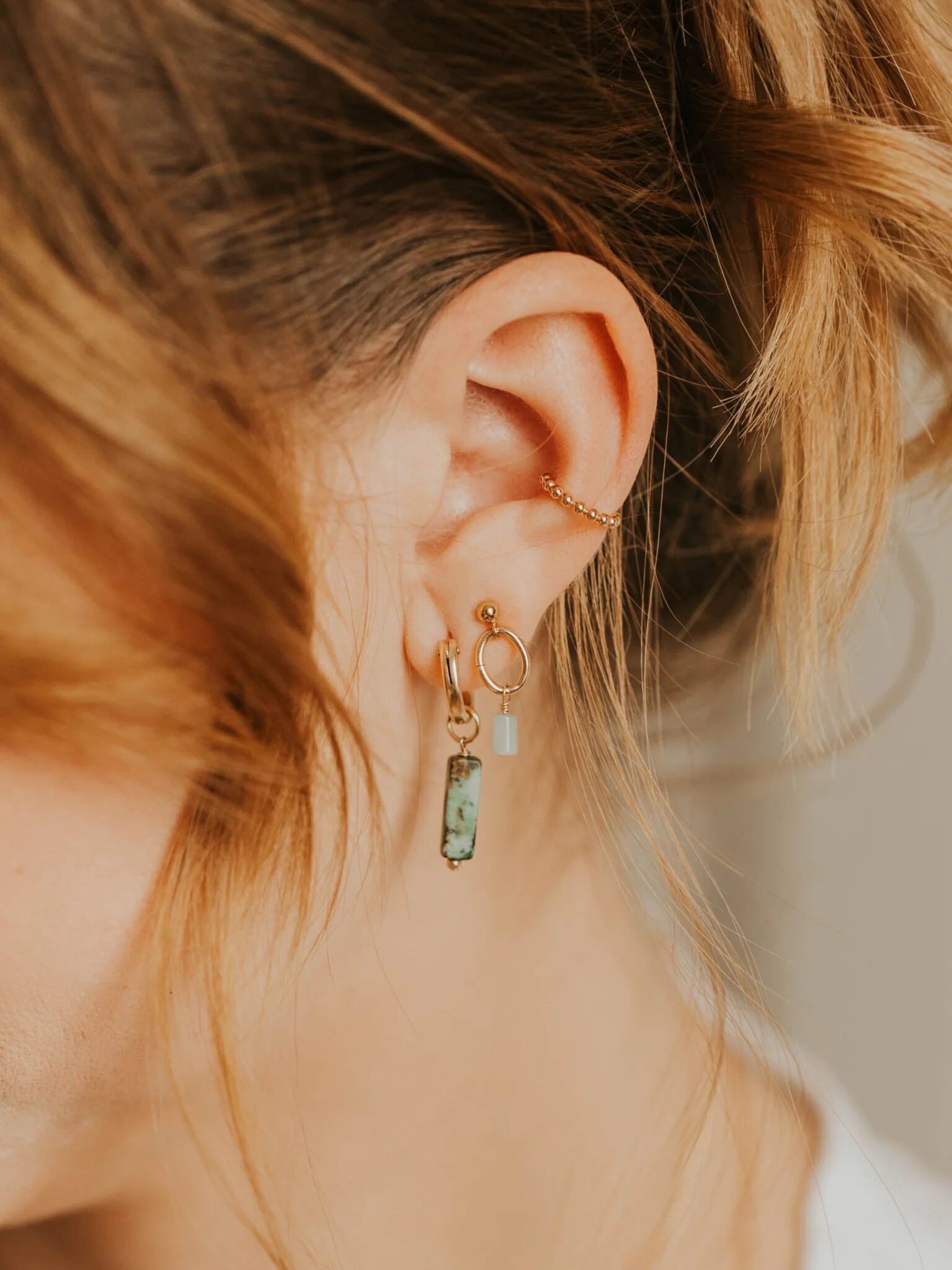 Close-up of a person's ear with a gold ear cuff and three small, gold hoop earrings. Two earrings feature turquoise accents. The person has light brown hair tied back loosely.