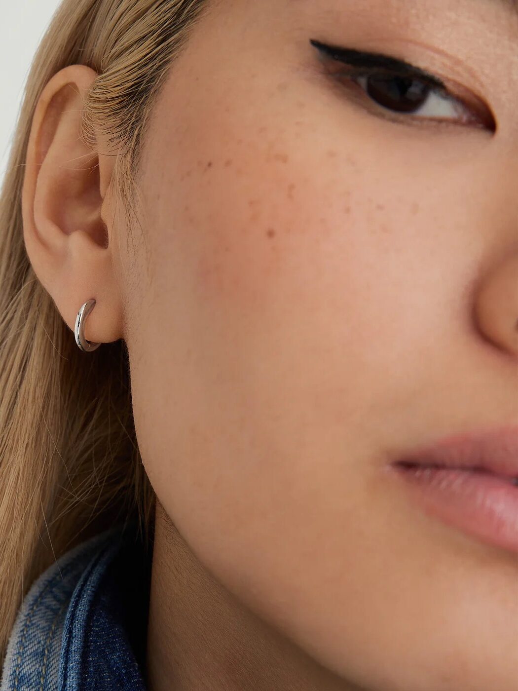 Close-up of a person with light blonde hair, showing a silver hoop earring and winged eyeliner. The person is wearing a denim jacket.