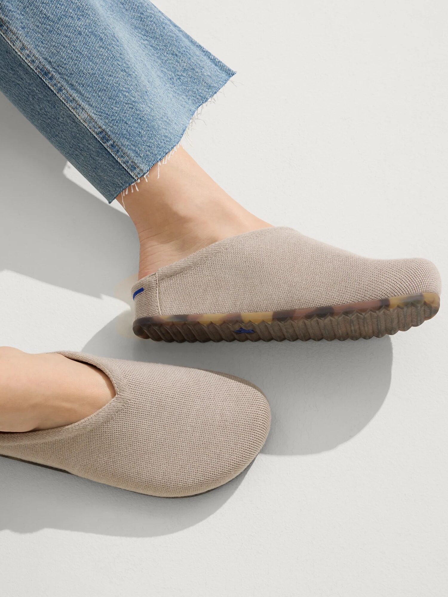 Person wearing frayed-hem blue jeans and beige slip-on shoes with textured soles on a white surface.