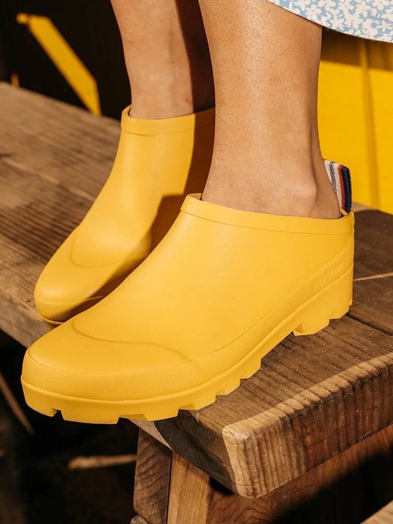 Person wearing yellow slip-on waterproof boots stands on wooden steps near a bright yellow wall.