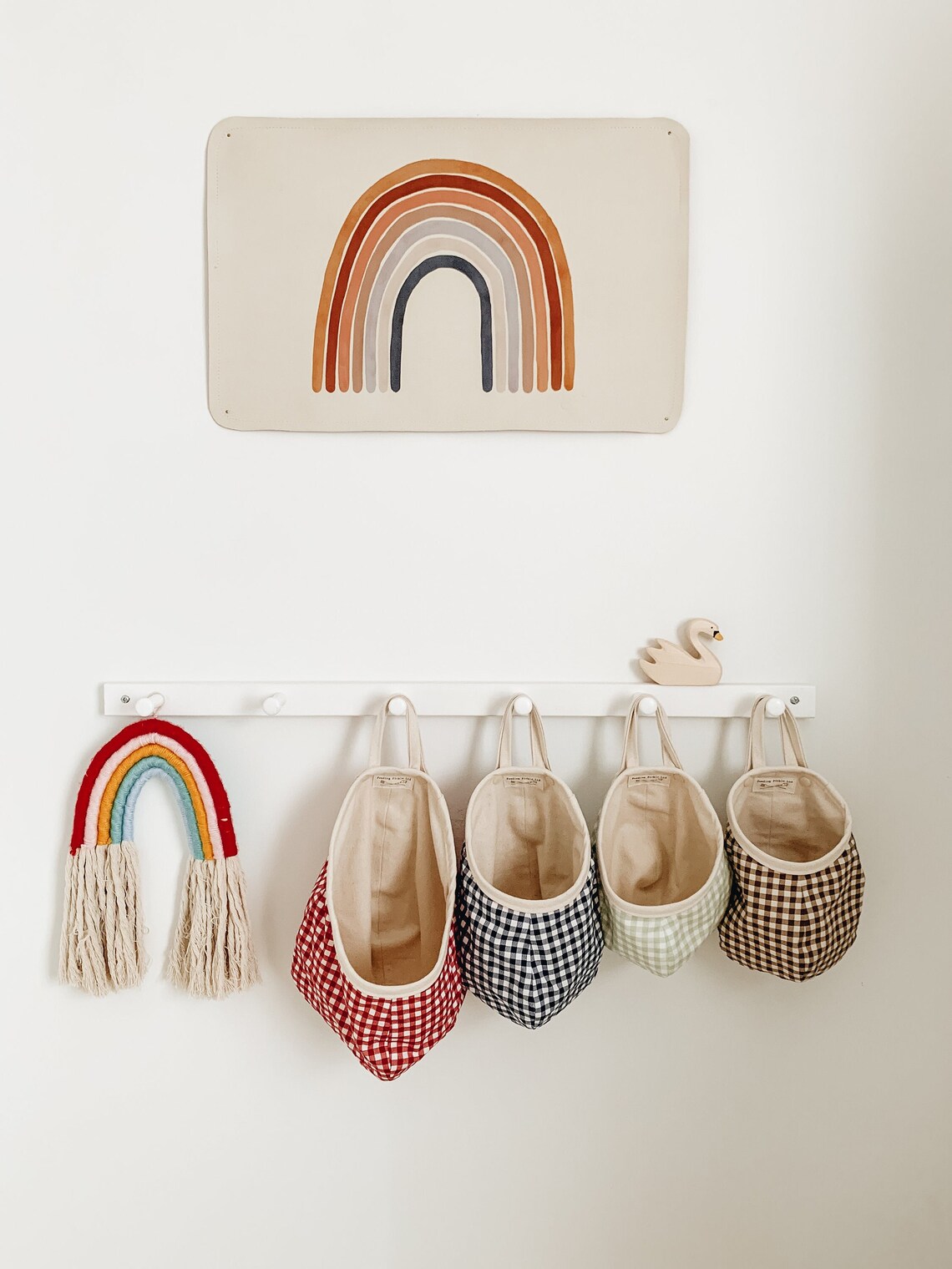 A wall with a rainbow art piece, a rainbow tassel, four gingham fabric bins hanging on hooks, and a small swan decoration on a white peg rail.