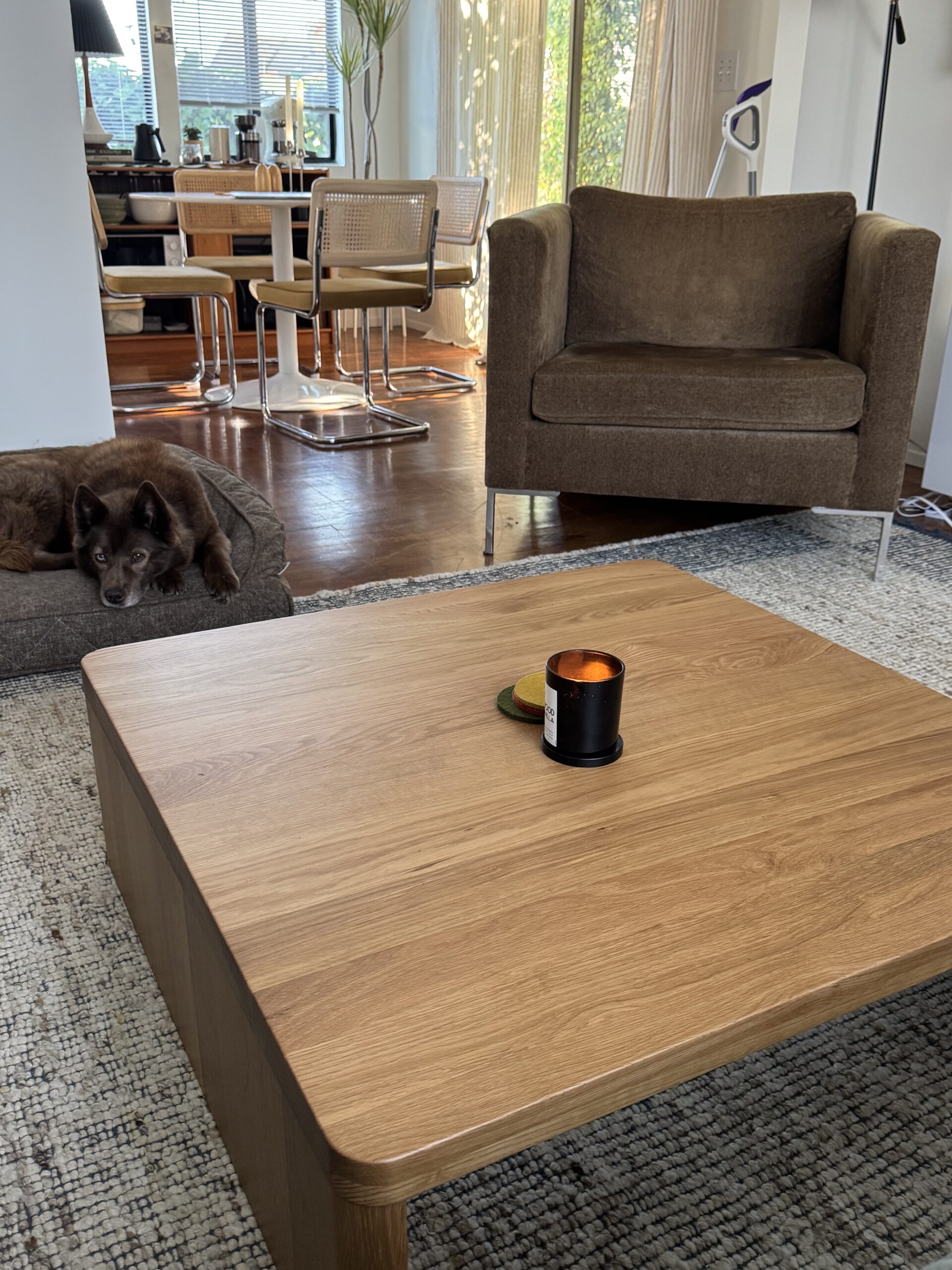 A lit candle sits on a wooden coffee table in a living room with a brown chair, a dog on a pet bed, and a dining area in the background.