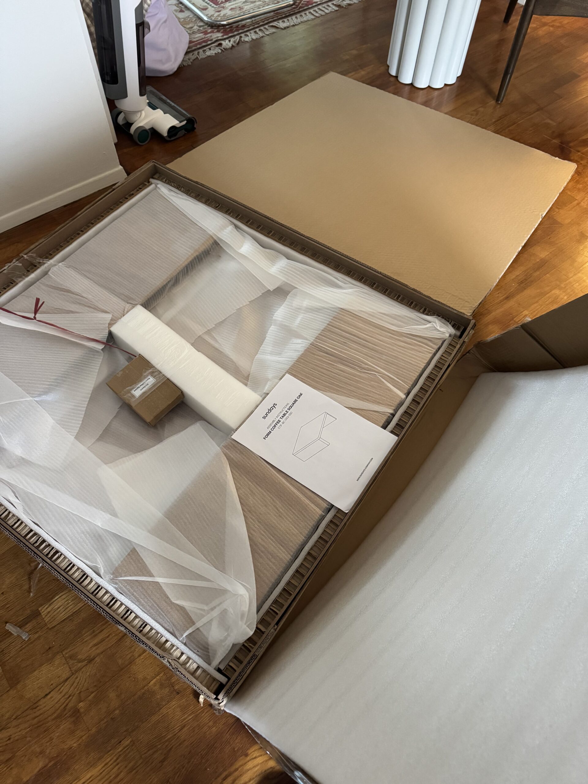 A flat-pack furniture kit with parts, instructions, and packaging materials laid out on a wooden floor, ready for assembly.