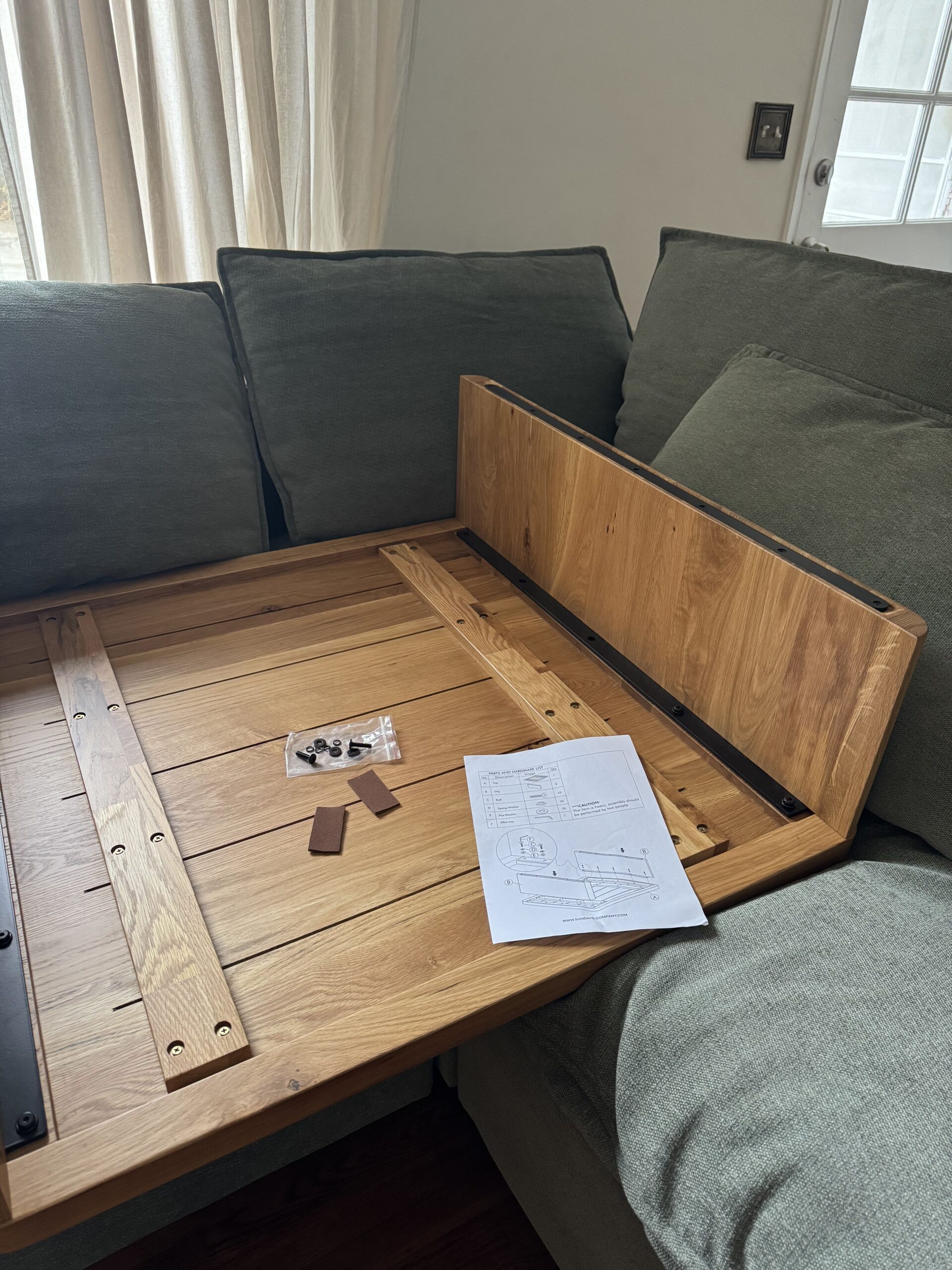 A partially assembled wooden table rests on a green sofa, with assembly instructions, screws, and small parts laid out nearby.