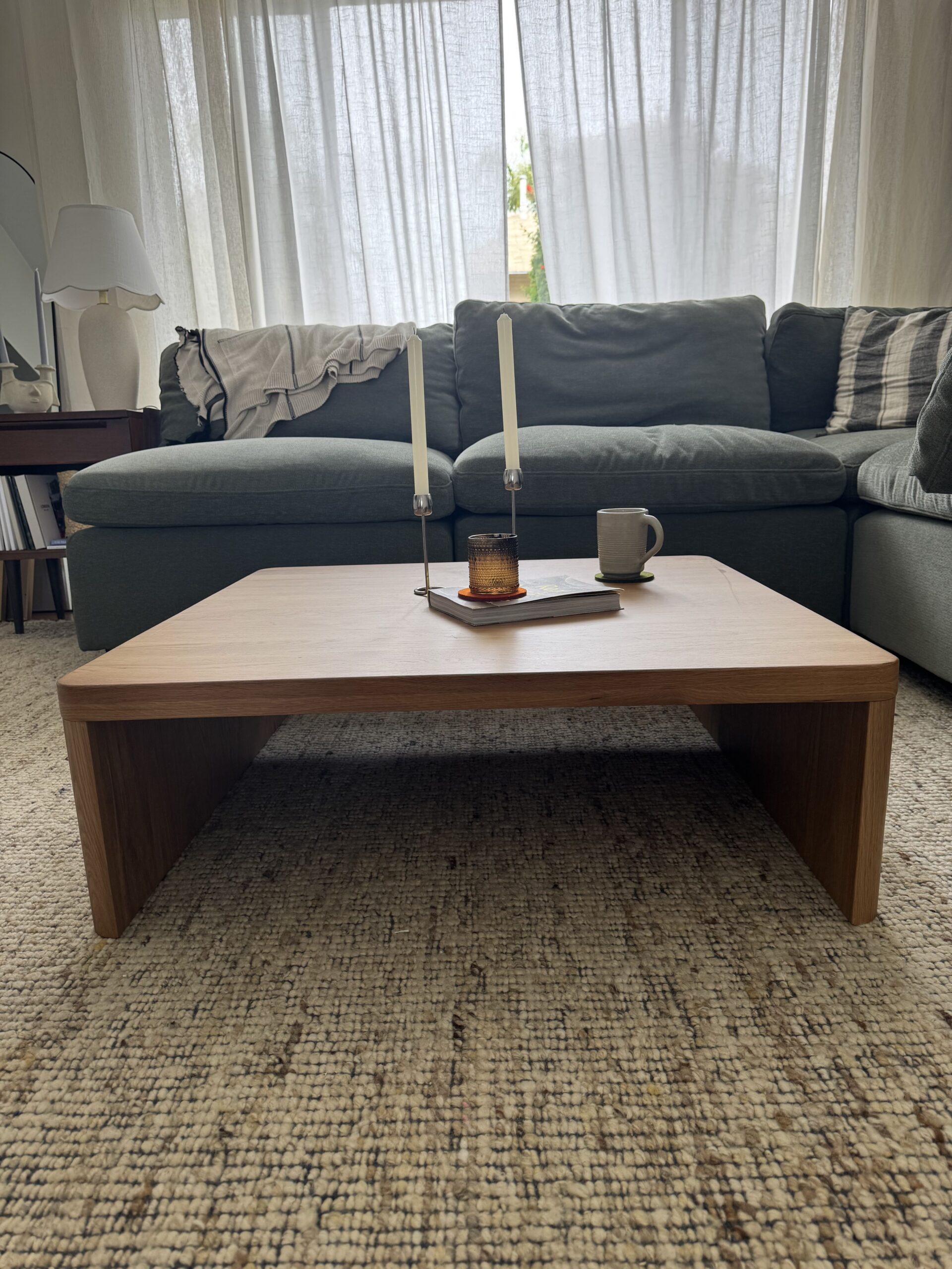 A wooden coffee table with two candles, a brown glass holder, and a mug, set in front of a green sectional sofa in a living room with light curtains.
