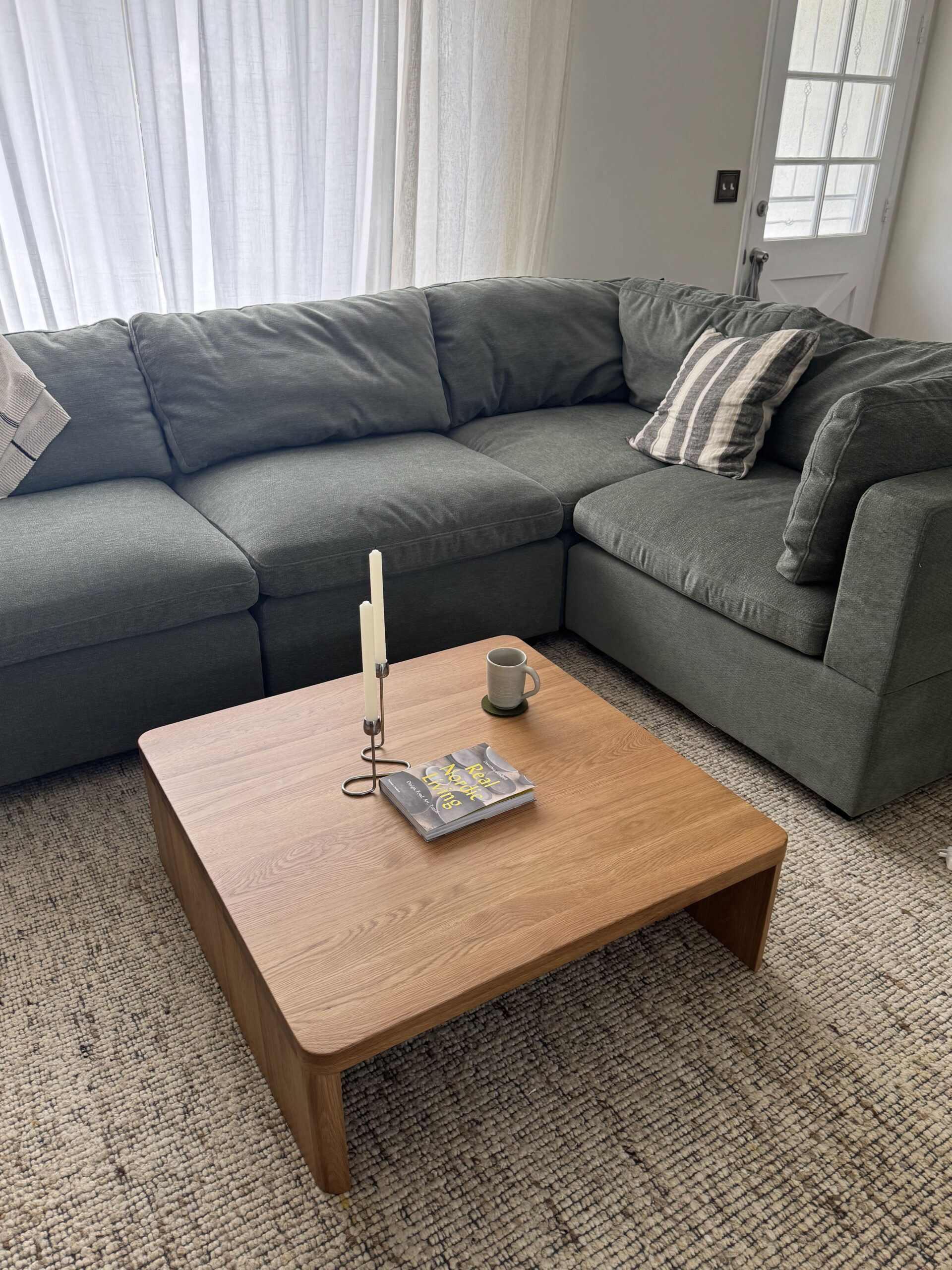 A green sectional sofa with striped pillows is next to a wooden coffee table holding a candle, a mug, and a magazine in a bright living room.