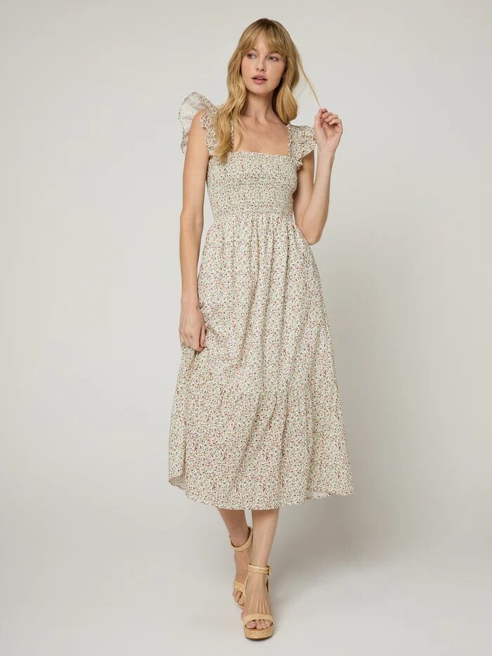 A woman stands wearing a light-colored, floral-patterned, sleeveless midi dress with ruffled shoulder straps and beige platform sandals against a plain background.