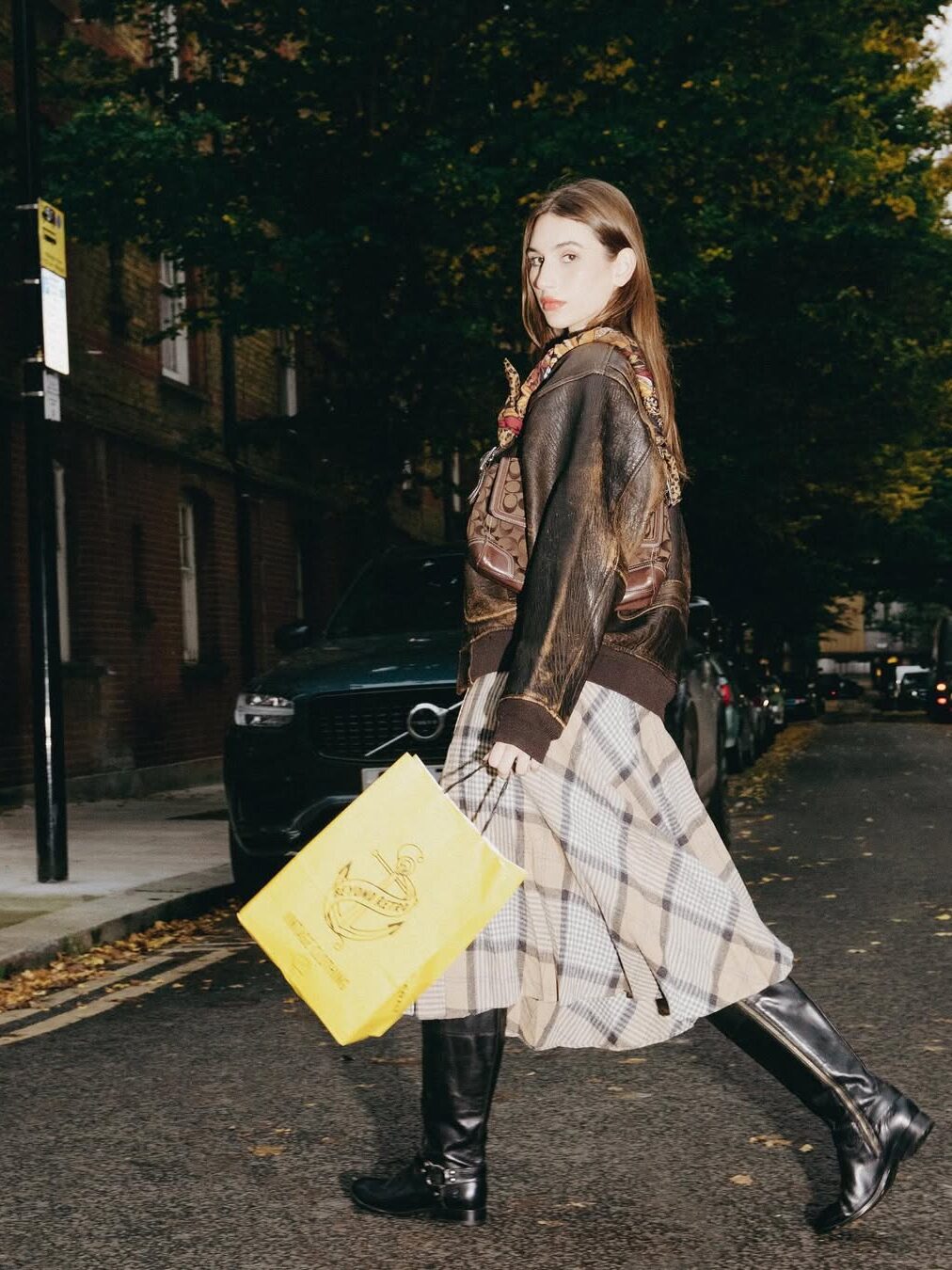 A woman in a brown leather jacket, plaid skirt, and black boots walks across a street holding a large yellow shopping bag. Trees and parked cars line the street.