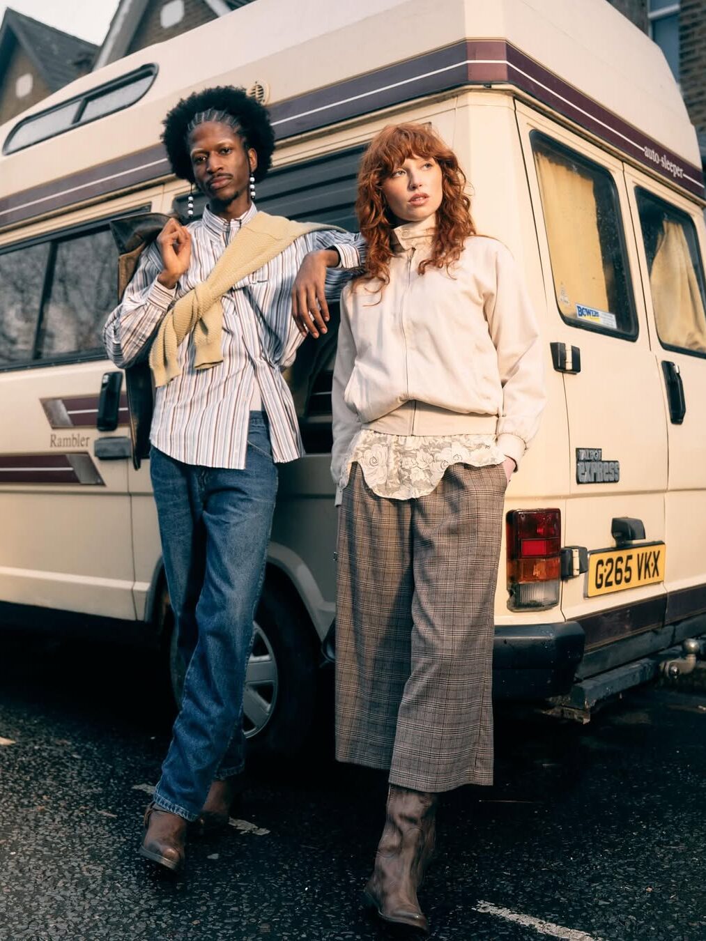 Two people stand in front of a cream and brown camper van parked on a street. One leans against the van, and both wear casual, vintage-inspired clothing.