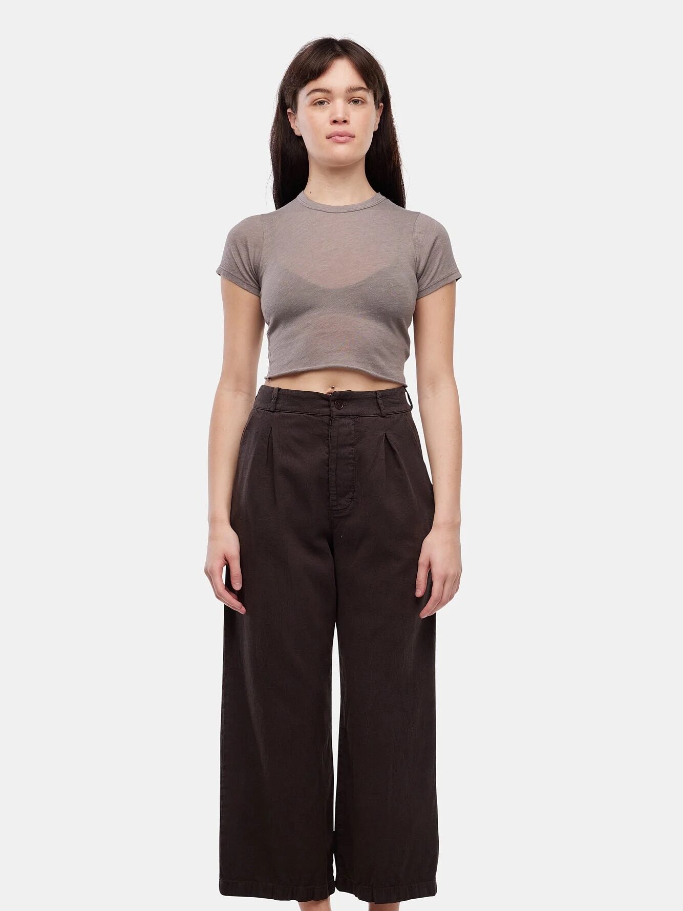 A woman stands facing forward wearing a short-sleeved, gray crop top, high-waisted dark wide-leg pants, and black flip-flops against a plain white background.