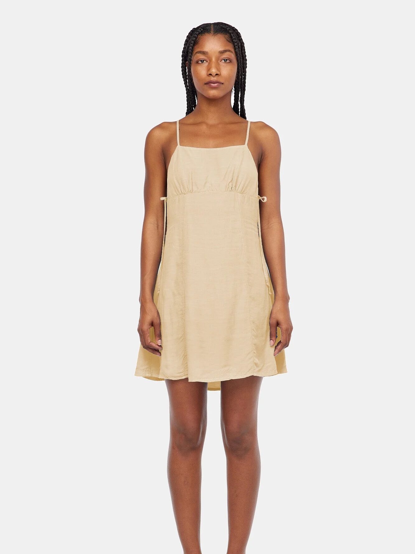 A woman stands facing forward wearing a sleeveless, light beige, knee-length dress with thin straps and black sandals against a plain white background.