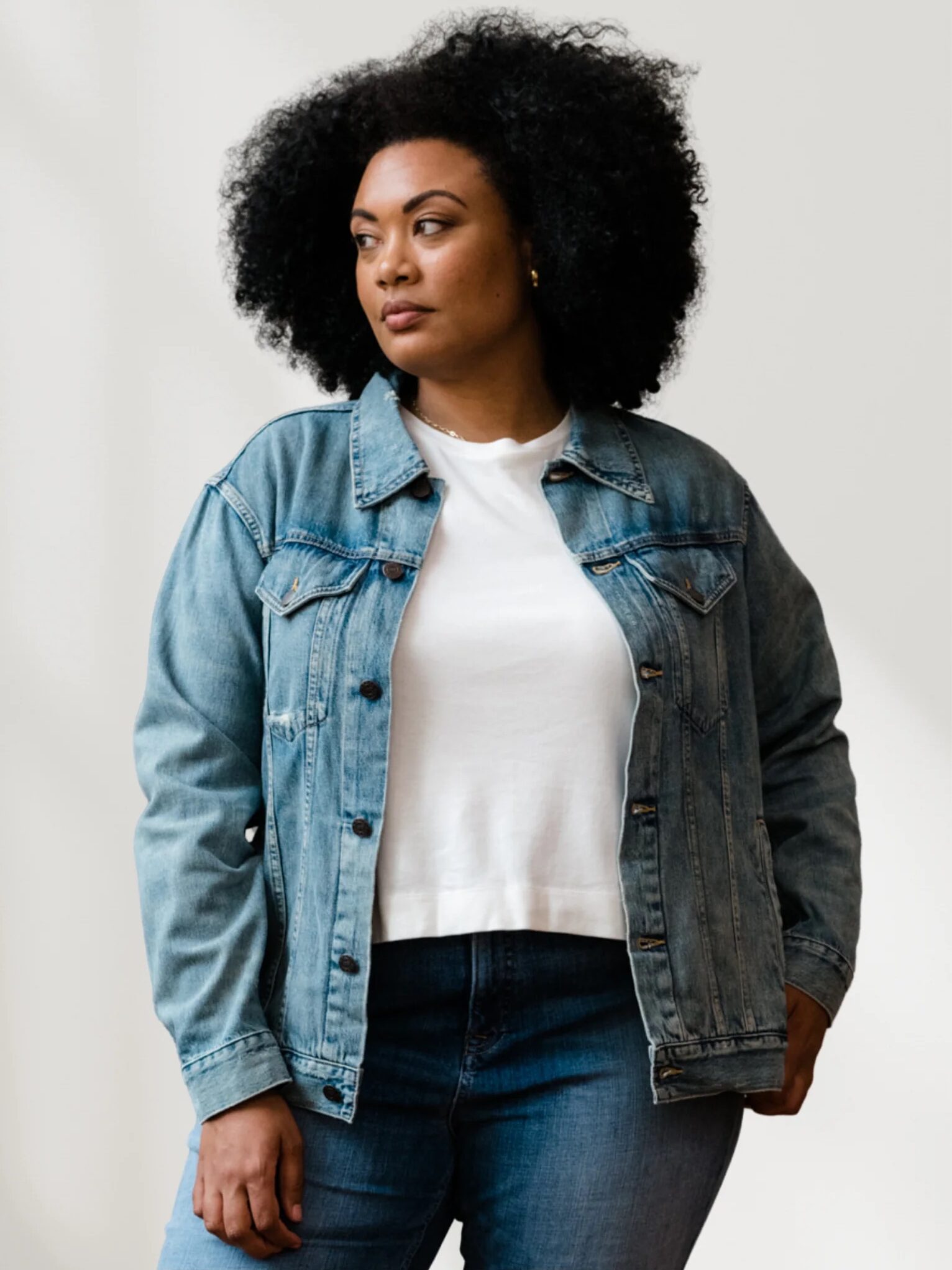 A woman stands against a plain background wearing a blue denim jacket over a white t-shirt and blue jeans, looking slightly to the side.