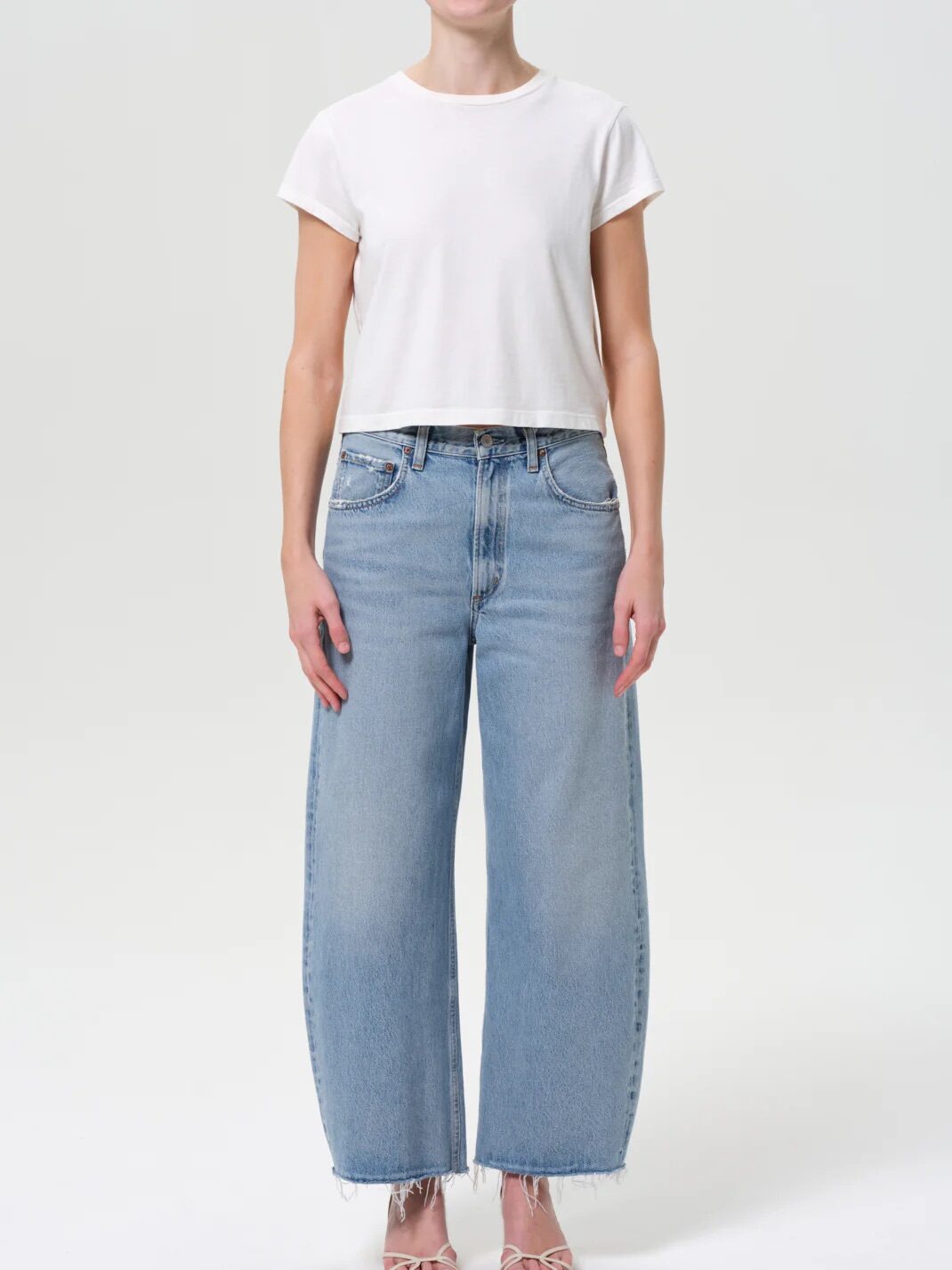 A person wearing a plain white short-sleeve crop top, light blue wide-leg jeans with frayed hems, and open-toe sandals stands against a white background.