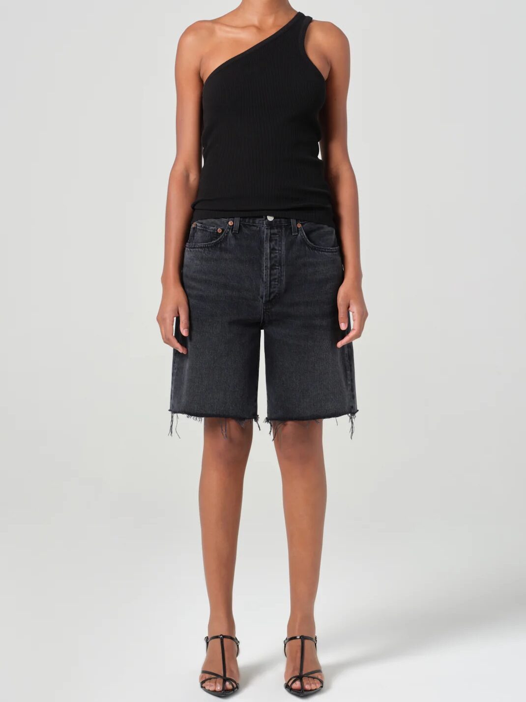 A person wearing a black one-shoulder top, black knee-length frayed denim shorts, and black strappy heels stands facing forward against a plain background.