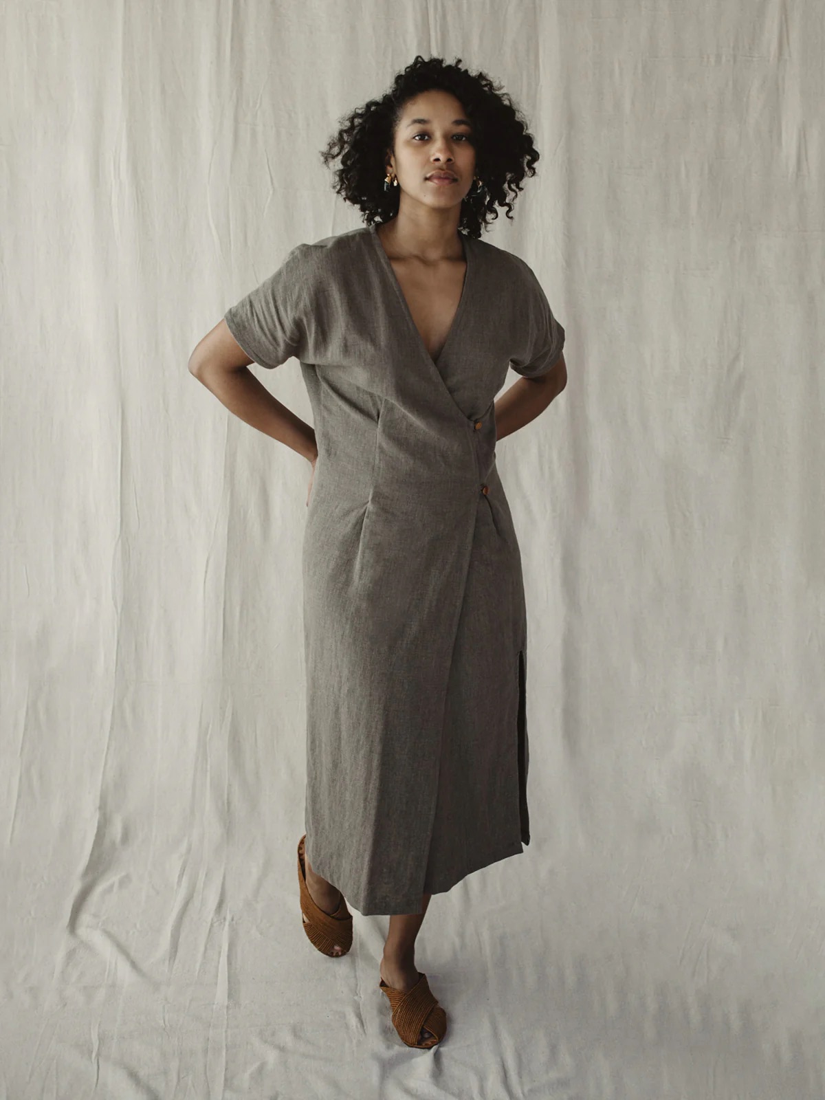 A woman stands against a neutral backdrop wearing a grey wrap dress and brown heeled sandals, with her hands behind her back.