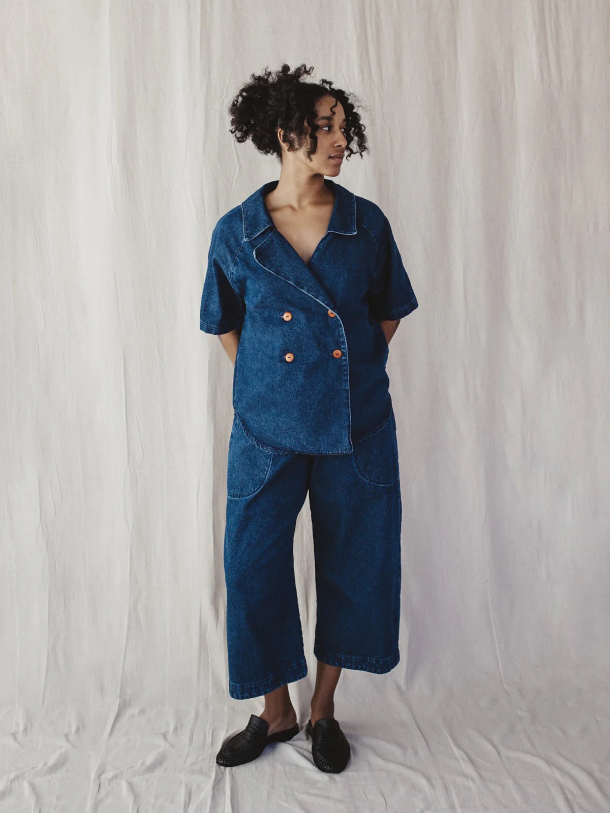 Person standing against a light fabric backdrop, wearing a blue denim short-sleeve jacket with orange buttons, matching wide-leg denim pants, and black slip-on shoes.