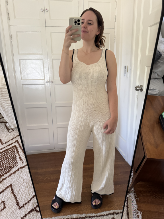 Woman taking a mirror selfie wearing a sleeveless, wide-leg cream knit jumpsuit and black sandals in a room with white cabinets and wooden floor.