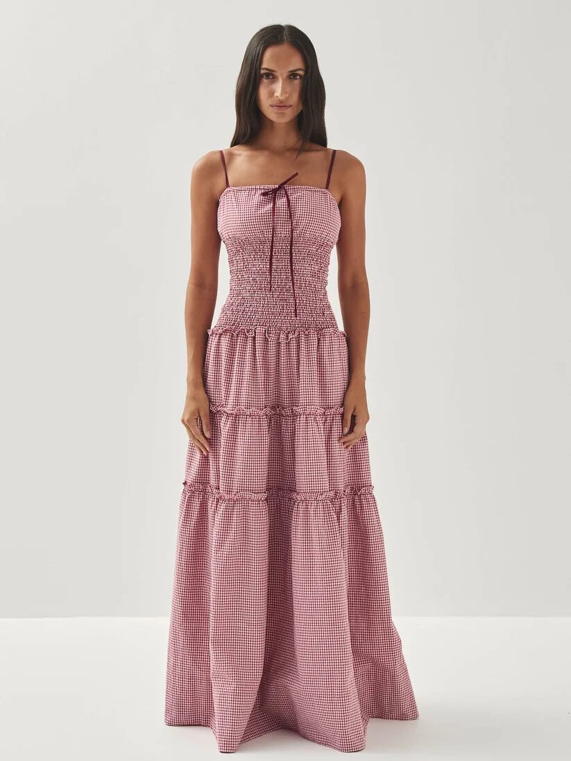 Woman standing against a plain background wearing a long, pink gingham dress with a fitted bodice, spaghetti straps, and tiered skirt.