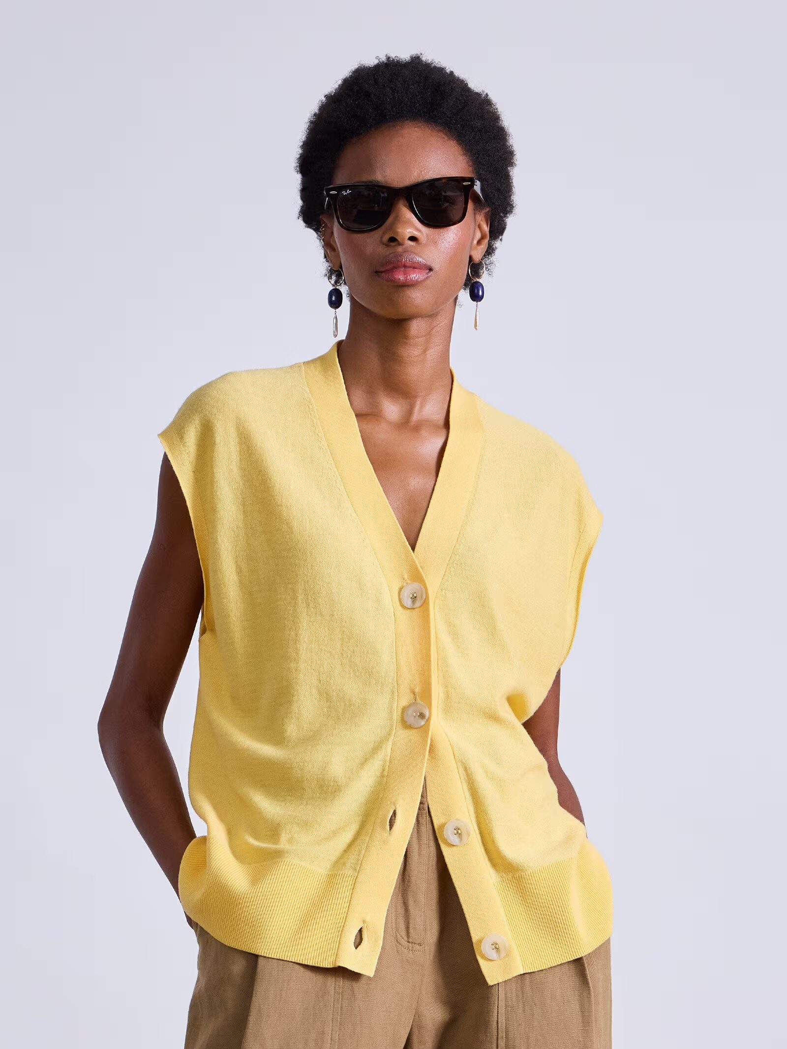 A person wearing sunglasses, a sleeveless yellow button-up vest, and light brown pants stands with hands in pockets against a plain background.