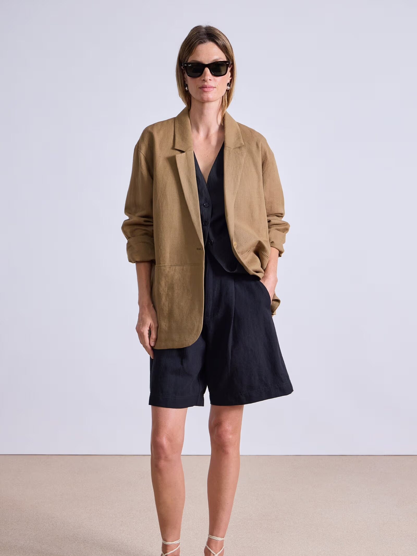A woman wearing sunglasses, a tan blazer, black shirt, black shorts, and brown sandals stands against a plain background with one hand in her pocket.