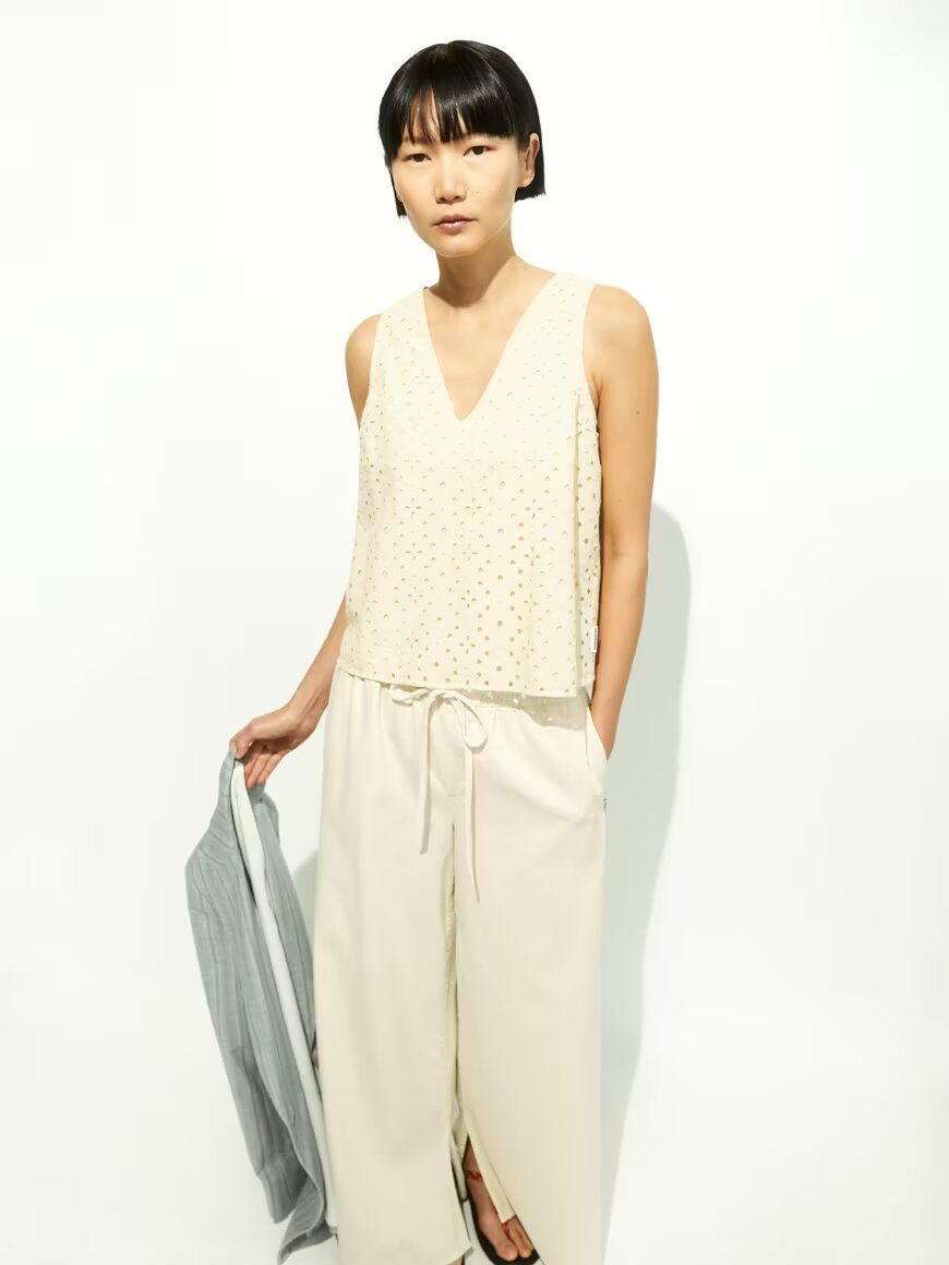 A person stands against a white background wearing a sleeveless beige top and wide-leg cream pants, holding a light grey garment in one hand.