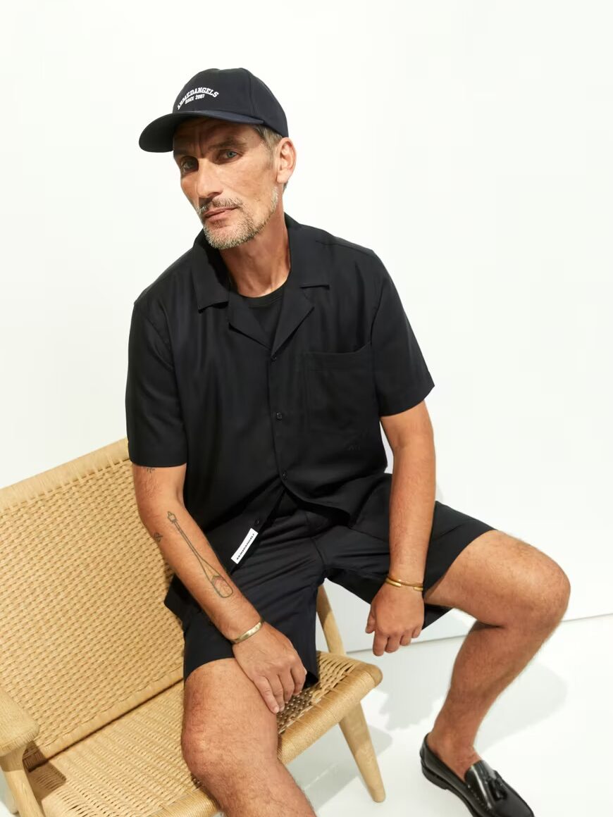 A man in a black button-up shirt, black shorts, black cap, and black loafers sits on a woven chair against a plain white background.