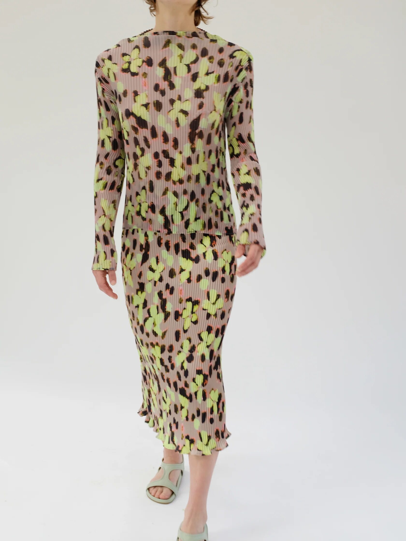 Person wearing a long-sleeved, midi-length dress with a green, pink, and black abstract pattern, and light green open-toe sandals, standing against a plain white background.