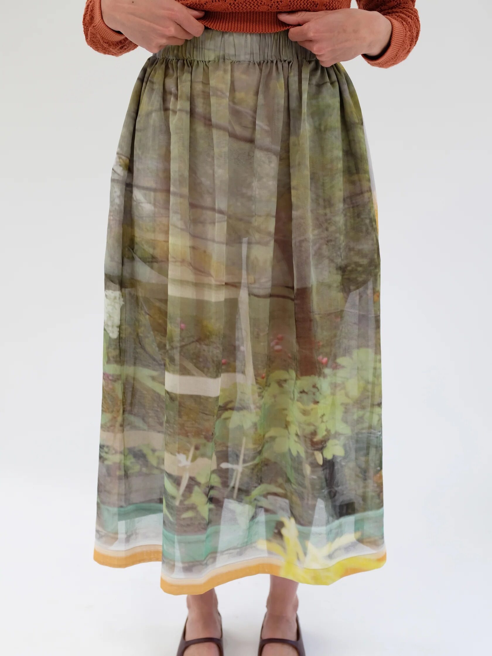 Person wearing a sheer skirt with a nature print, an orange top, and brown shoes, standing against a plain white background.