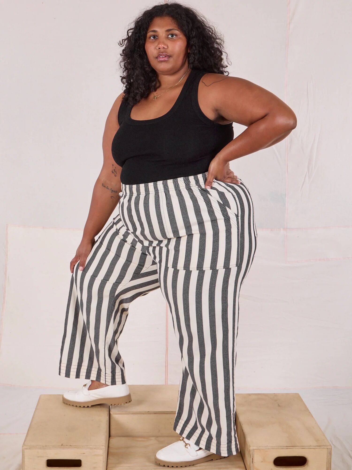 A person with curly hair wearing a black sleeveless top, black and white vertically striped pants, and white shoes stands on wooden platforms against a plain backdrop.