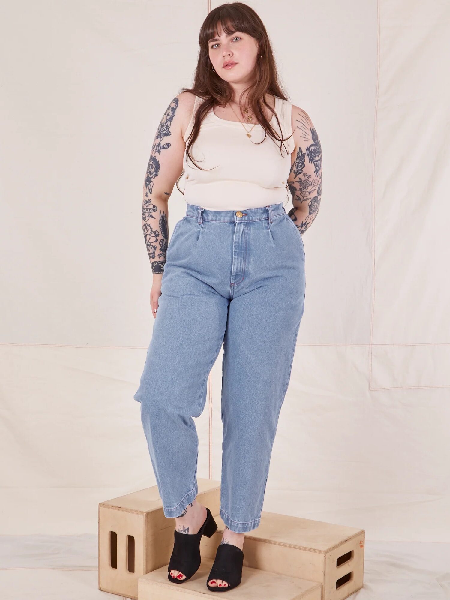 Woman with long brown hair and tattoos wears a white tank top, high-waisted blue jeans, and black sandals, standing on wooden boxes against a plain light backdrop.