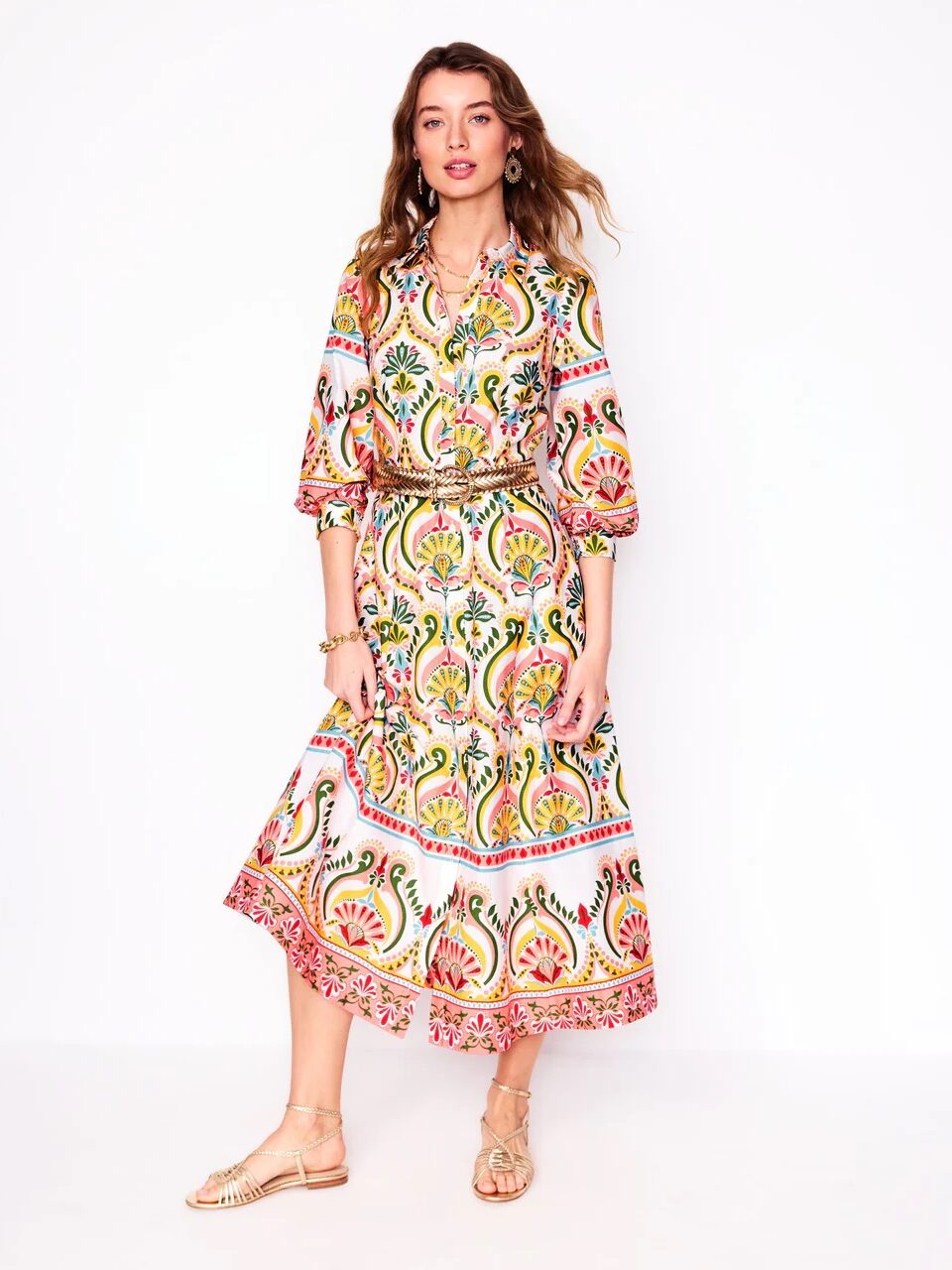 Woman standing against a white background, wearing a colorful patterned dress with long sleeves and a midi skirt, paired with beige sandals and jewelry.