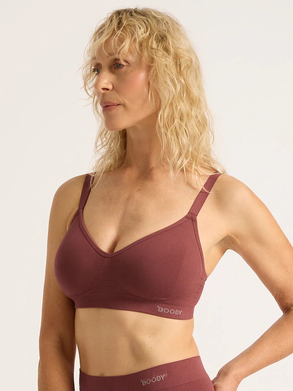 A woman with shoulder-length blonde hair wears a maroon wireless bra and matching bottoms, standing against a plain background.