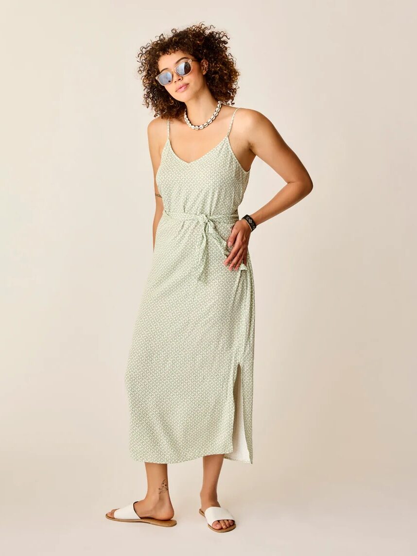 A woman stands against a plain background wearing sunglasses, a light green sleeveless midi dress with a belt, white slide sandals, and a beaded necklace.