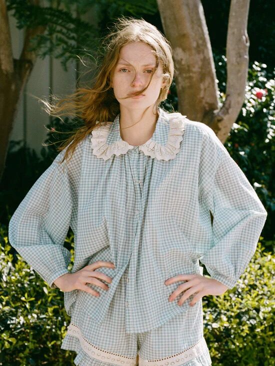 A person stands outdoors wearing a loose, light blue gingham pajama set with white lace trim on the collar and shorts.