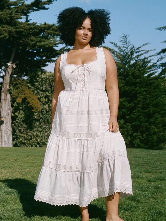 A woman stands barefoot on grass wearing a sleeveless, white, tiered summer dress with lace details; trees and blue sky are in the background.