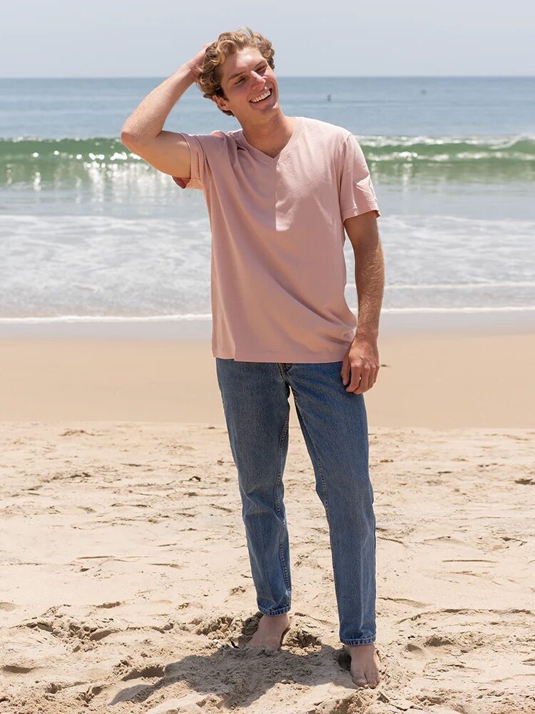 A man stands barefoot on a sandy beach, wearing a light pink T-shirt and blue jeans, with one hand touching his head. The ocean and sky are visible in the background.