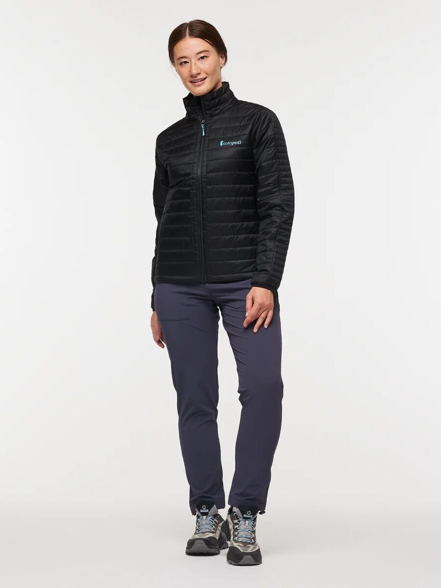 A person stands facing forward, wearing a black zip-up jacket, dark pants, and hiking shoes against a plain white background.