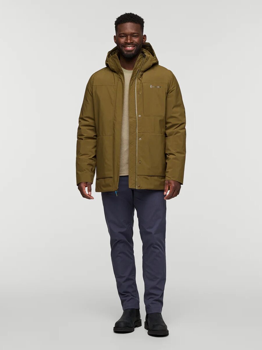 A man stands facing forward wearing an olive green hooded parka, beige sweater, dark blue pants, and black shoes against a plain light background.