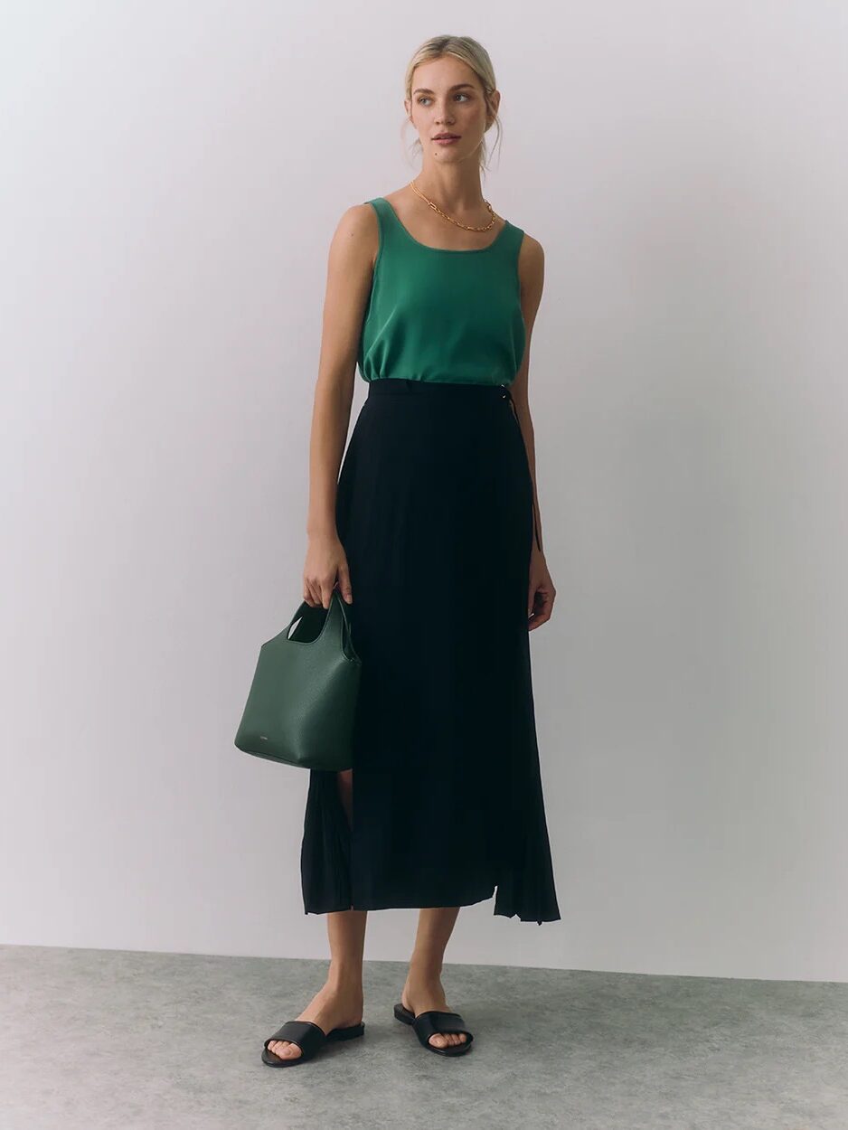 A woman stands against a plain background wearing a green sleeveless top, black midi skirt, black sandals, and holding a small green handbag.