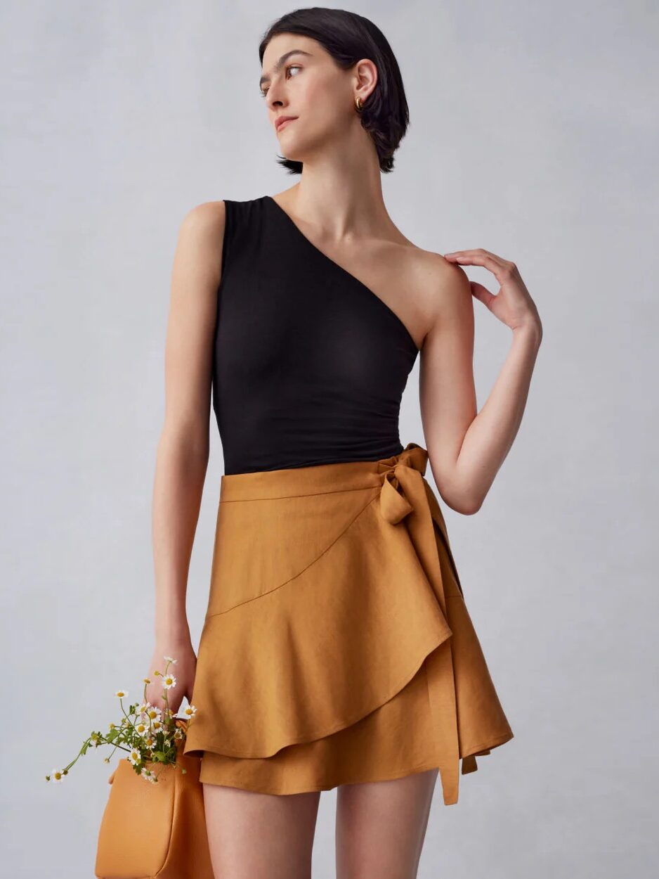 A woman wearing a black one-shoulder top and a mustard yellow wrap skirt stands holding a small handbag with flowers against a plain background.