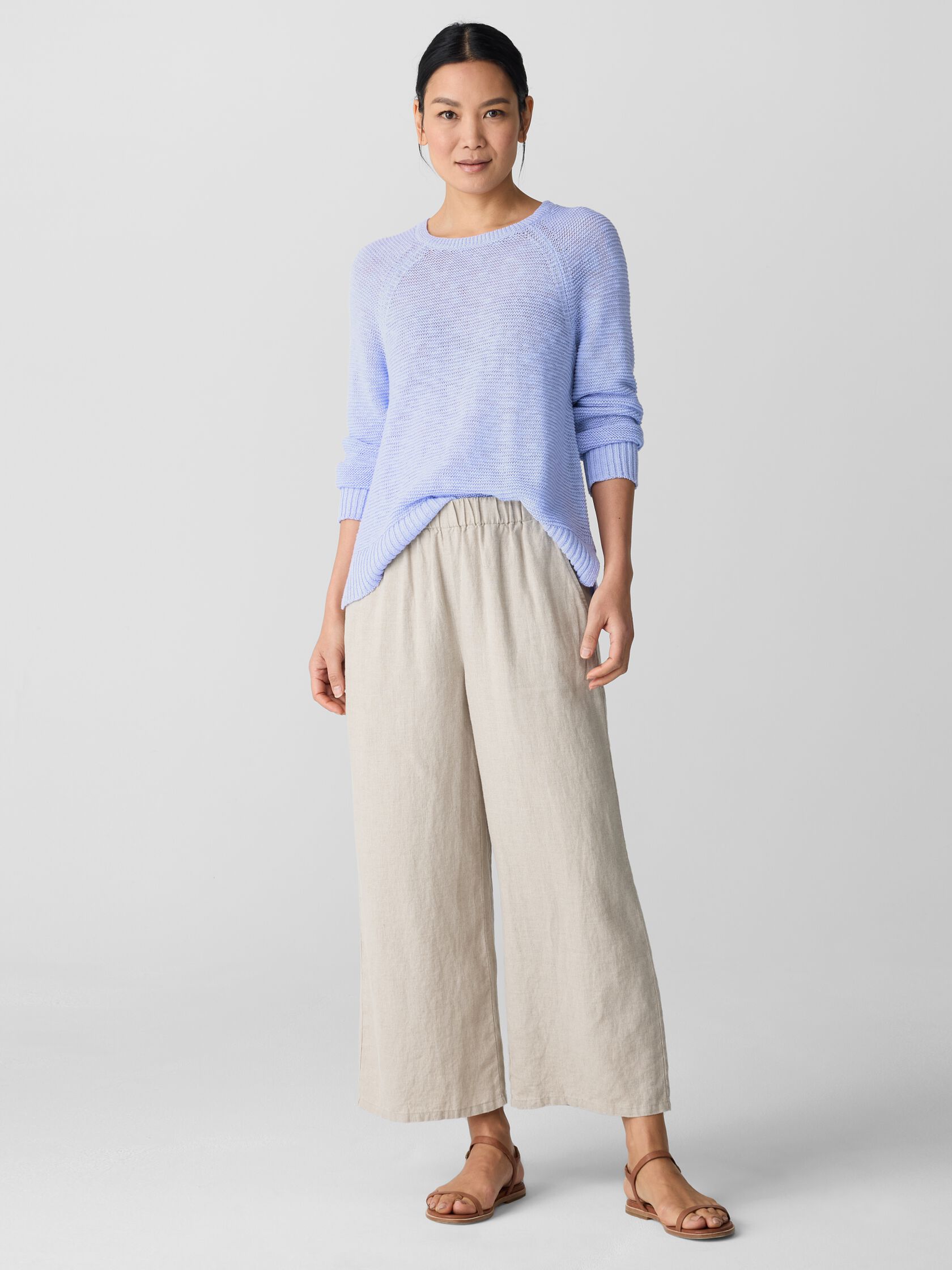 A woman stands in a light blue sweater, beige wide-leg pants, and brown sandals against a plain light background.