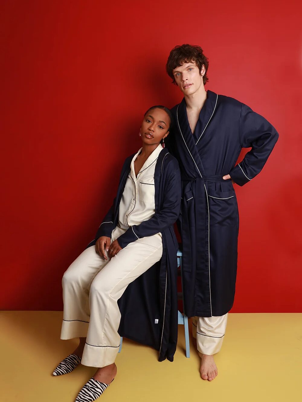 Two people wearing matching navy robes over cream pajamas pose against a red background; one is seated, the other stands with hand on hip.
