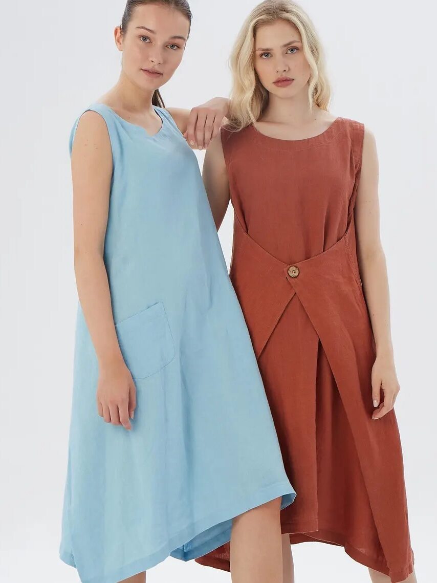 Two women stand side by side wearing sleeveless linen dresses; one in light blue with a large front pocket, the other in rust with a cross-over front detail and a button.