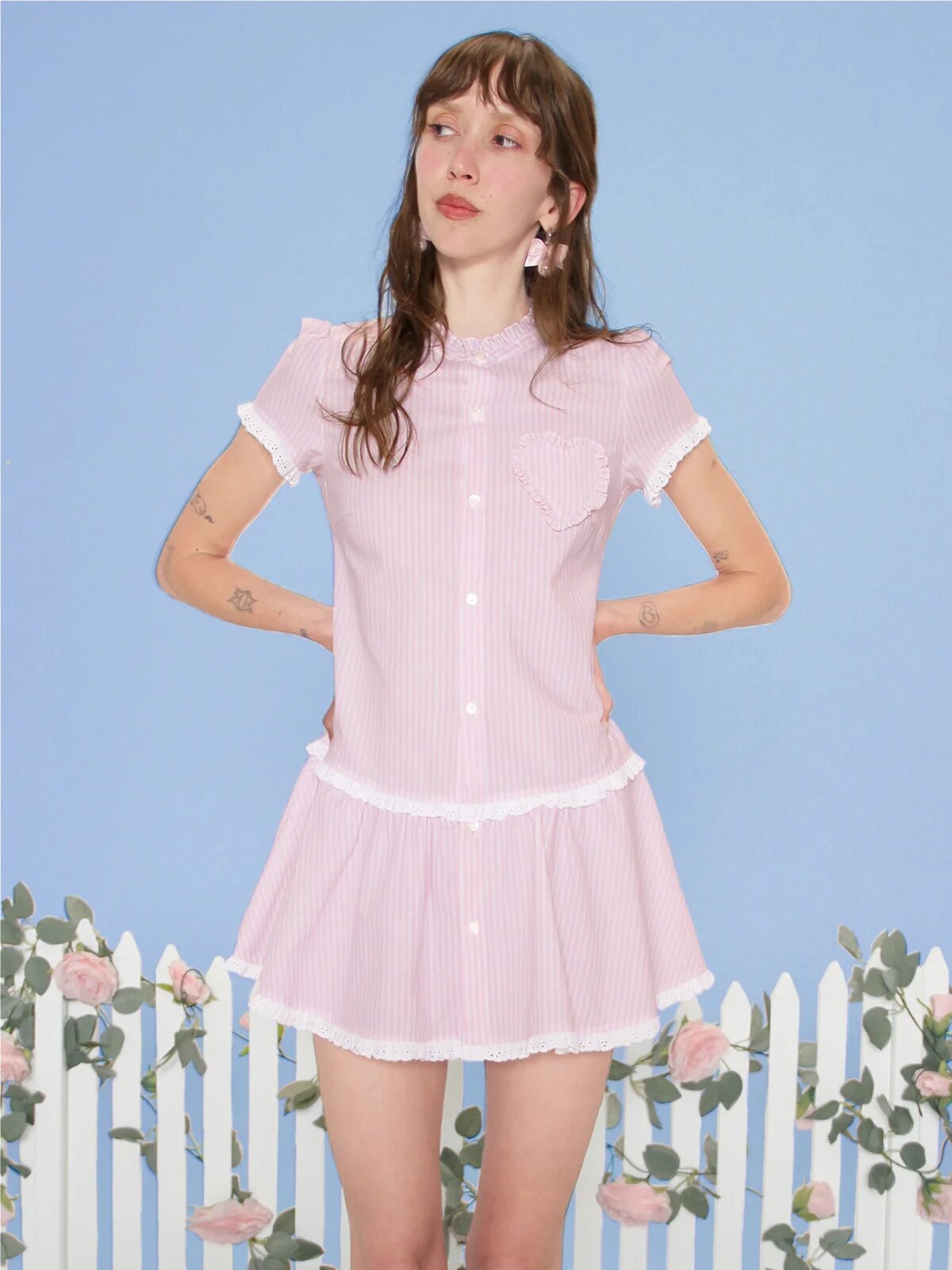 A person stands in front of a white picket fence with roses, wearing a pink and white short-sleeve dress with a ruffled skirt and knee-high socks, against a blue background.