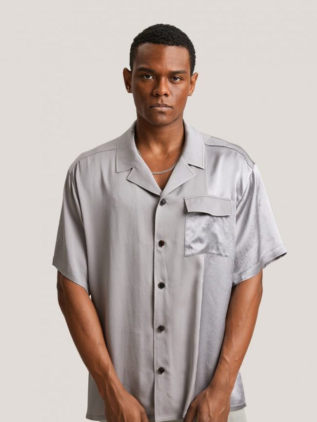 A man wearing a short-sleeve, light gray satin button-up shirt with a chest pocket, standing against a plain light background.