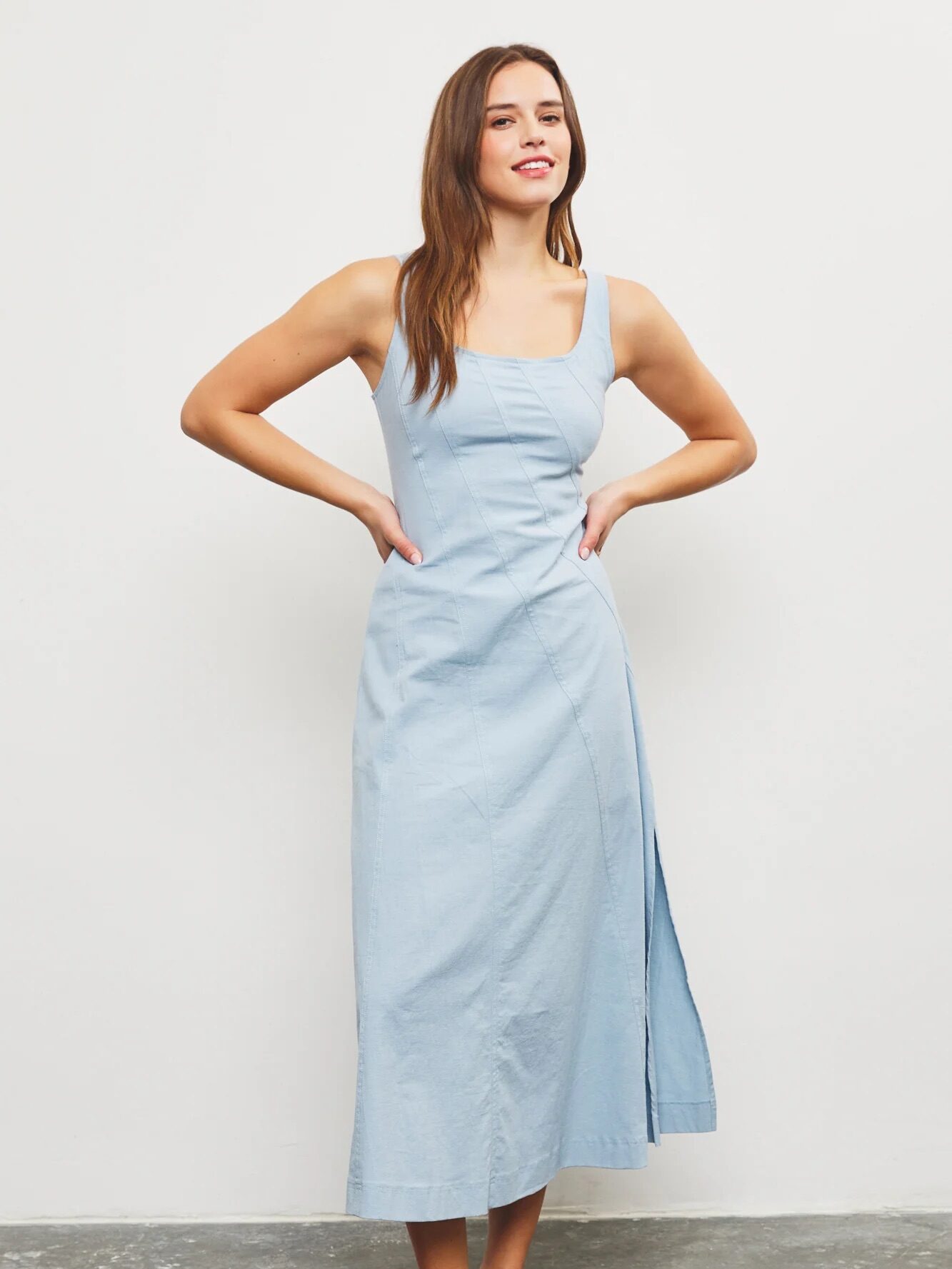 A woman stands against a plain wall wearing a light blue sleeveless midi dress with a square neckline and side slit, and black slide sandals with decorative embellishments.