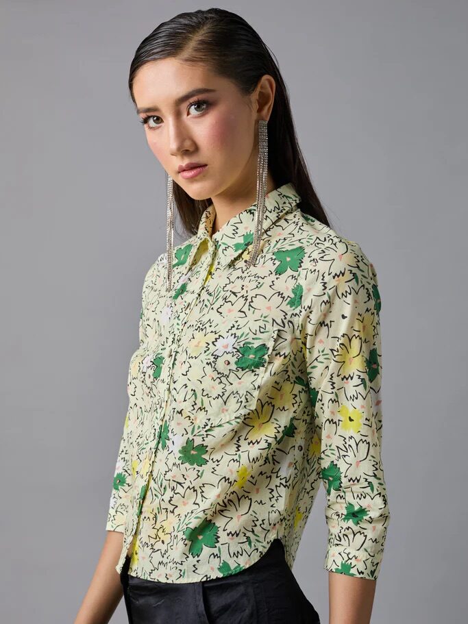 A woman stands against a plain grey background wearing a floral button-up shirt with green and yellow patterns and black pants, with long straight hair and dangling earrings.